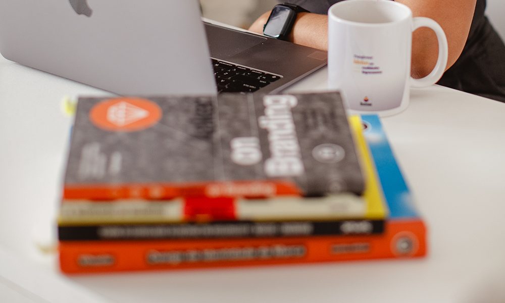 Livros de branding e design empilhados ao lado de um notebook e uma caneca da Pastana